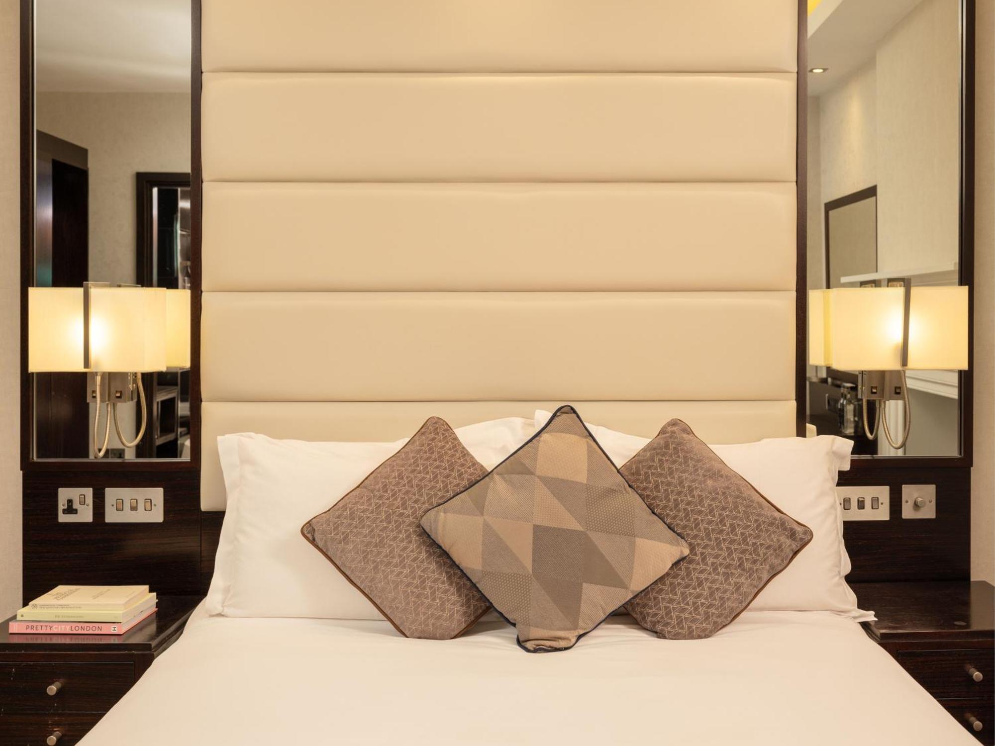 Montcalm Brewery Townhouse Hotel London Bagian luar foto The image shows a cozy bedroom setting. The bed features a headboard upholstered in a light beige fabric. It is adorned with three decorative pillows in varying patterns and colors, placed on white bedding. On either side of the bed, there are modern