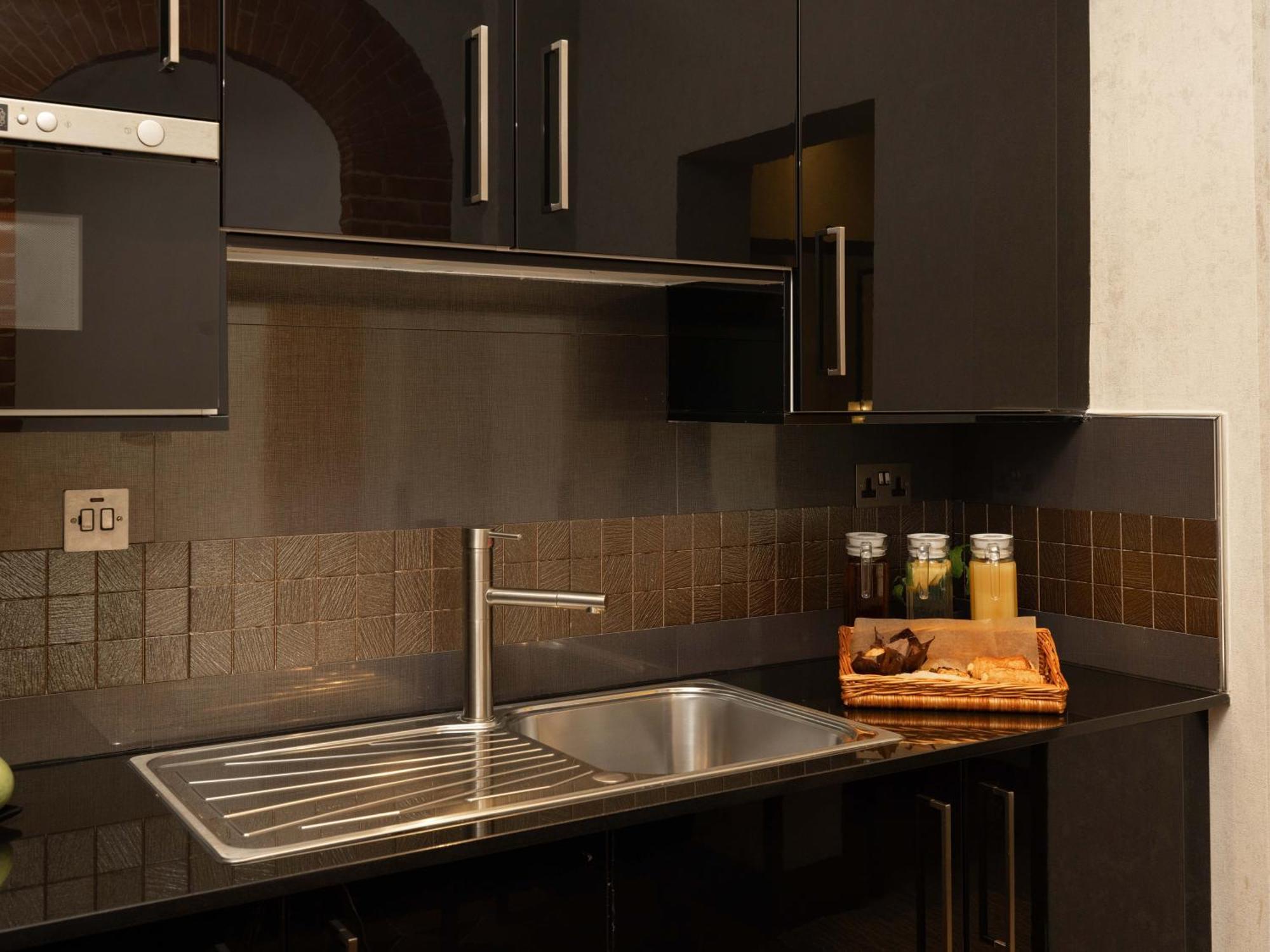 Montcalm Brewery Townhouse Hotel London Bagian luar foto The photo shows a modern kitchen with a sleek design. It features dark cabinetry with a glossy finish and stainless steel handles. There is a stainless steel sink with a simple faucet, and a countertop made of black material. Above the sink, there ar