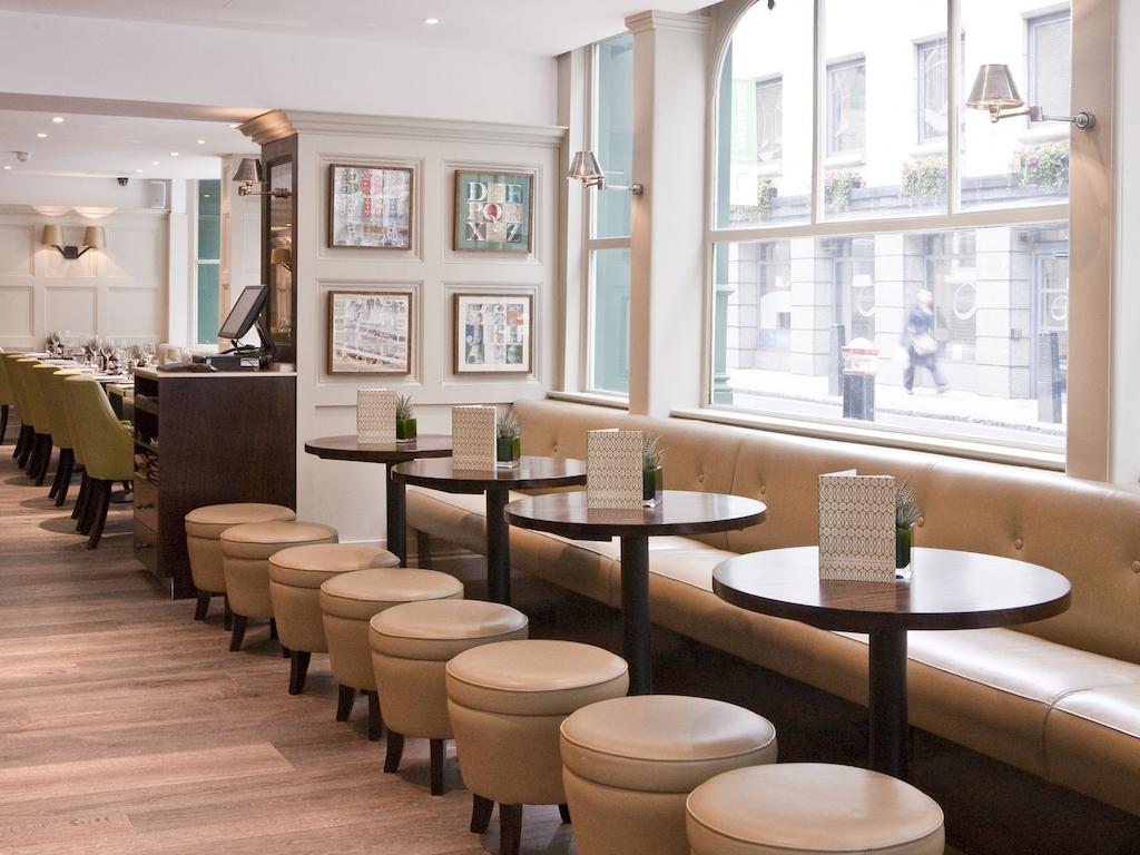 Montcalm Brewery Townhouse Hotel London Bagian luar foto The photo displays a modern café or restaurant interior. There is a cozy seating area featuring light-colored, upholstered benches along the wall, accompanied by small round tables. The tables are paired with circular stools. The decor includes frame