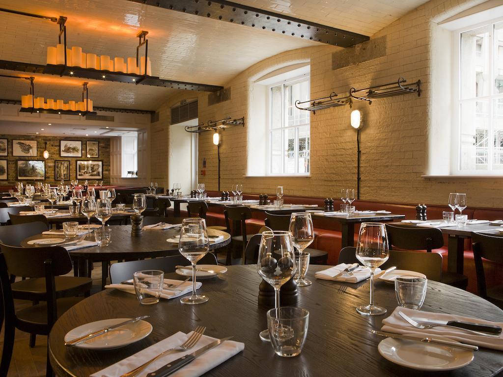 Montcalm Brewery Townhouse Hotel London Bagian luar foto The photo shows a restaurant interior featuring a warm and inviting atmosphere. It has several round wooden tables set for dining, each with neatly arranged plates, utensils, and glasses. The lighting is soft, with overhead fixtures providing a cozy 