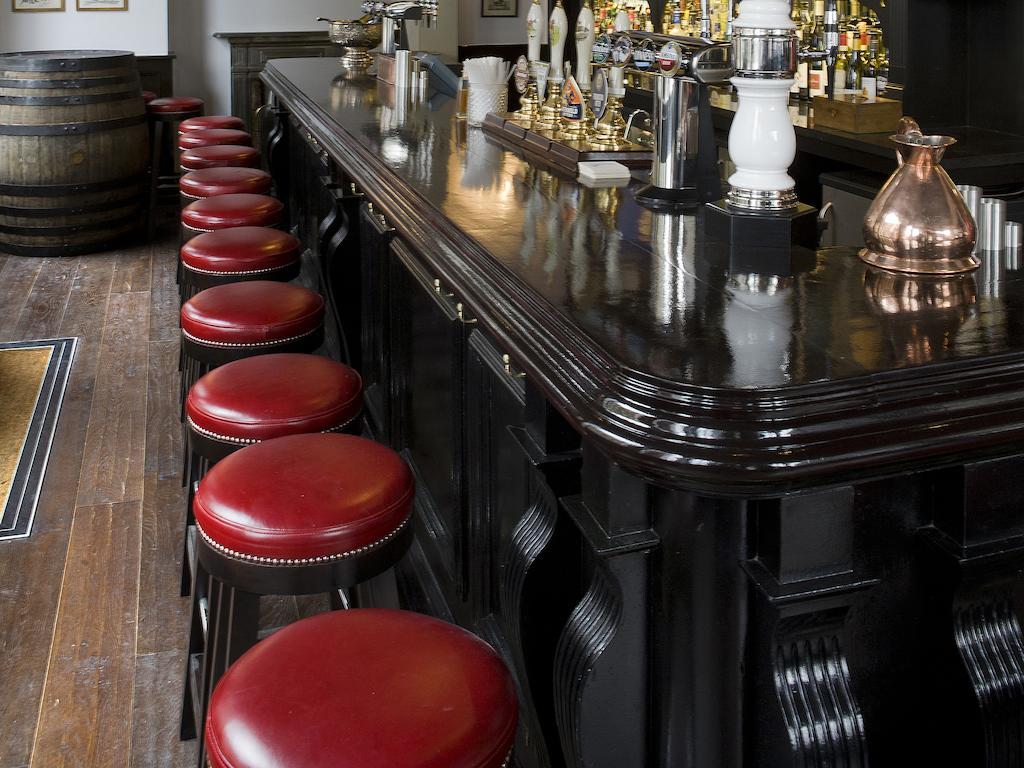Montcalm Brewery Townhouse Hotel London Bagian luar foto The photo shows a bar setting with a long, polished dark wooden bar counter. Attached to the counter are several bar stools with red leather seats. Behind the bar, there are various bottles, suggesting a well-stocked bar. The flooring appears to be w